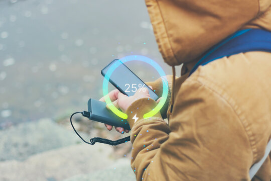 A traveler in the mountains charges his Smartphone with portable Power Bank. The battery indicator shows the percentage of charge. The concept of travel and outdoor chargers. - Powered by Adobe