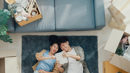 Top view. Happy Asian couple lay down on carpet use tablet shopping online furniture from decoration store in living room at new house. Husband wife move house, Apartment Rent And Ownership Concept.