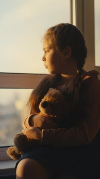 Sad little girl hugging teddy bear and looking through window, feeling lonely