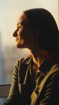 Cheerful red-haired woman looking through window, enjoying city sunset view