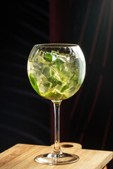 Refreshing green cocktail with mint leaves and ice cubes served in a large glass on a wooden board against a dark moody background