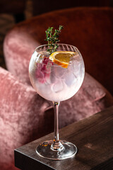 Elegant gin cocktail with ice, orange slice, thyme sprig, and berries served in a large glass on a wooden table with a soft velvet chair in the background