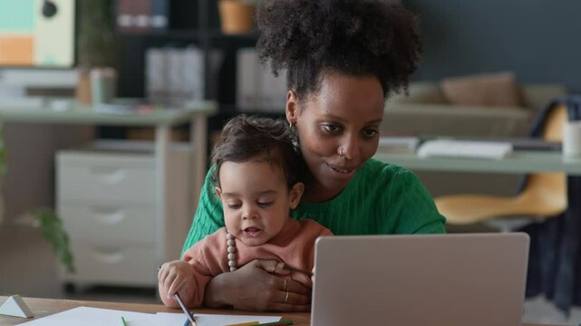 Medium shot of cheerful young woman babysitting small son while having online call on wireless laptop at office desk