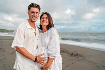 Obraz premium Young family posing for camera on the beach. Pregnant happy woman with her lovely husband touching gently her belly. Healthy relationships