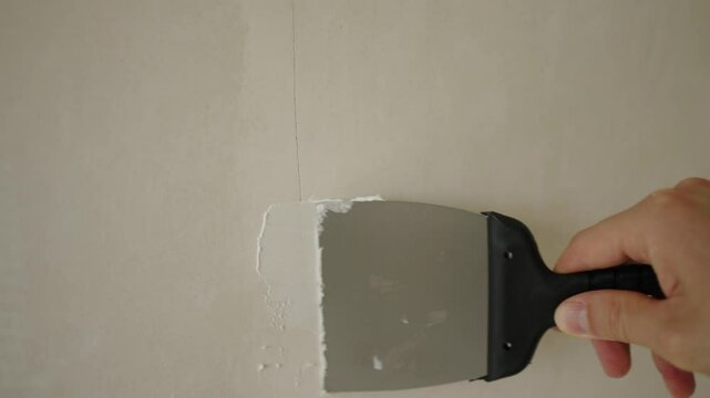 Construction professional carefully spreading putty across white wall surface, methodically filling crack and preparing wall section for upcoming painting project