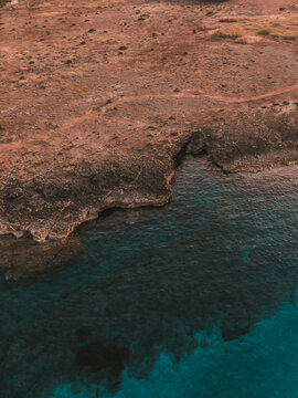 Aerial view of a rugged coastline where dry land meets vibrant turquoise waters. A visually dramatic contrast of textures and colors in a Mediterranean-like setting.