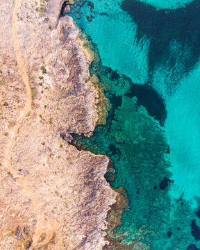 Dramatic aerial of rocky coastline with clear turquoise waters meeting the rugged land. A beautiful contrast of textures and colors, ideal for nature, travel, and coastal themes.
