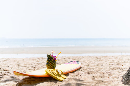 Beautiful decorated pineapple and flip-flops and surfboard left on the beach. Holidays, vacations, and summer vibes concept.