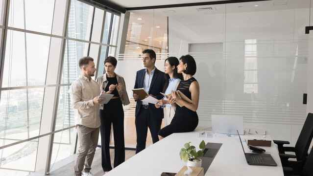 Five serious focused teammates gathered in skyscraper office meeting room for joint project discussion, listen European team member explain point, share new ideas and creative solution during briefing