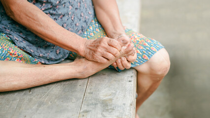 Elderly person sitting on wooden bench, holding foot with both hands, wearing patterned skirt and...