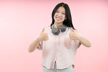 young asian woman giving thumbs up while wearing headphone in pink studio background