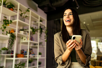 Young businesswoman smiling while using a mobile phone in a modern office filled with plants, radiating happiness and professionalism