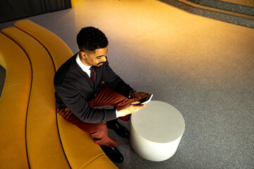 Young businessman sitting comfortably on a yellow sofa in a modern office lobby, using a smartphone to stay connected and engaged