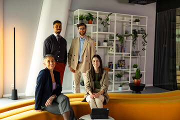 Four smiling business professionals posing together in a modern office filled with greenery and bookshelves, radiating teamwork and success
