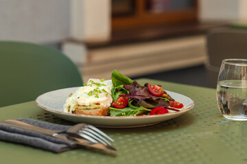 Delicious Eggs Benedict served with a side salad on a rustic plate for a perfect outdoor brunch on a summer day.Close-up of a gourmet brunch plate. Poached eggs on toast with fresh salad and cutlery