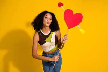 Stylish young woman holding a heart prop on vibrant yellow background, showcasing cheerful and...