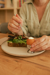 Close-up of female hands with cutlery having breakfast with egg and bacon with freshly greens served on wooden table in sunny outdoor cafe. Part of a series