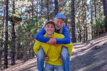Grandson carries his grandfather on shoulders during excursion day in forest. Smiling multi-generational family enjoying mountain and nature together. New generation care and helps the old one