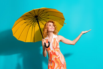 Attractive young woman holds a yellow umbrella against a teal background, wearing a stylish printed...