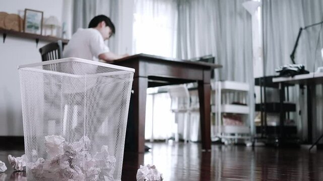 A man sitting at a desk tears and crumples a piece of paper before throwing it into a white aluminum trash can at home