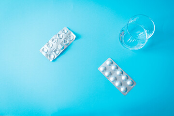 Blister packs with white pills and a glass of water on a blue background. Conceptual flat lay for medication, daily supplements, and treatment routine