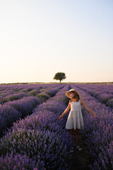 Little girl in summer dress and straw hat standing in lavender field at sunset. Peaceful childhood...