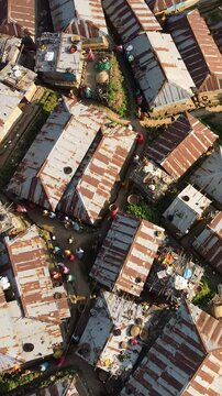 Aerial video of a sprawling low-income settlement with densely packed houses, rusted metal roofs, and winding dirt paths. Small figures, one in vibrant red, walk below. Natural warm daylight, deep
