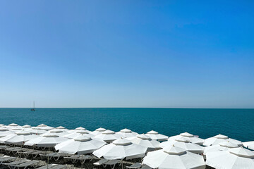 White beach umbrellas and white sun loungers on the shore of a bright blue sea on a cloudless summer day. Background, backdrop, screen, desktop.