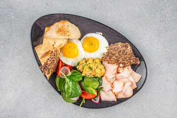 Healthy breakfast plate with sunny side eggs, avocado mash, greens, turkey slices, and toast.