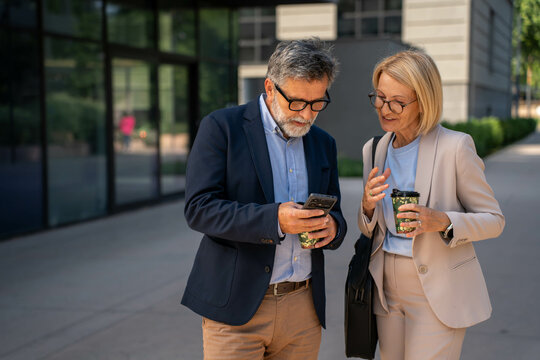 Diverse mature business professionals standing outdoors, chatting and looking at a smartphone