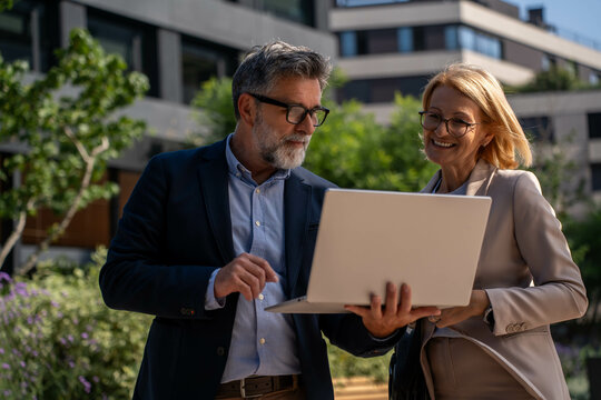 Successful Mature Businessman and Businesswoman with Laptop Outdoors, Happy and Confident in Beautiful Business District - Powered by Adobe