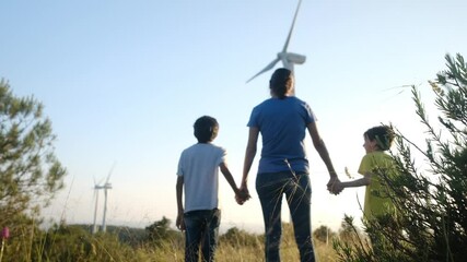 Mother holding hands with two children, standing together near wind turbines on verdant hillside, symbolizing family unity and renewable energy commitment