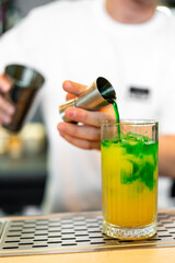 A bartender pours a green liquid into a glass with yellow liquid and ice, creating a vibrant layered cocktail. The close-up shot highlights the bartending process and drink preparation