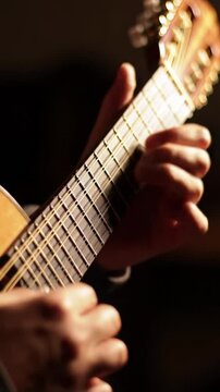 Tight close-up video of musician's hands in dark hoodie, skillfully strumming/picking strings of charango-like acoustic instrument against dark creamy bokeh background, illuminated by soft warm