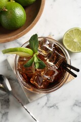 Tasty Cuba Libre cocktail in glass, limes and spoon on white marble table, flat lay