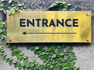 A brass entrance sign with a pointing arrow on a green covered wall
