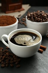 Cup of drink, ground coffee, beans and cinnamon on black table, closeup