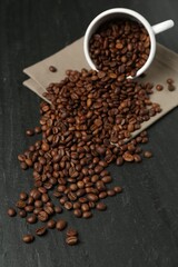 Aromatic coffee beans and overturned cup on black table, closeup