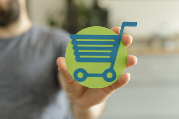 Close-up of a hand holding a paper cutout of a shopping cart, symbolizing online shopping or...