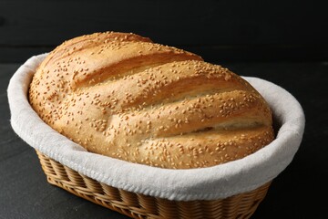 Loaf of bread with sesame seeds in basket on black table, closeup