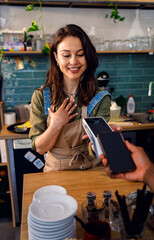Female barista charges the customer for takeaway coffee in cozy modern coffee shop.