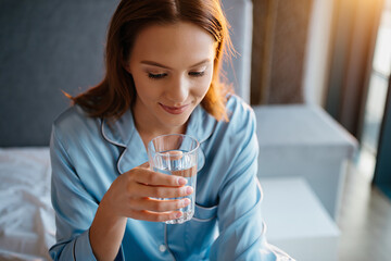 Young woman in blue pajamas drinking water in bed