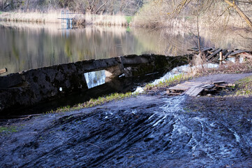 Lake with sinking concrete wall. Muddy ground with narrow tire tracks. Small stream crossed by wooden pallet bridge. Dry reeds on far shore, path continues beyond bridge.