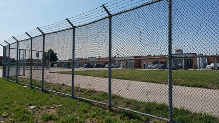 A fenced area with a chainlink fence and barbed wire running along the top, surrounded by green grass and a paved surface in the foreground, with buildings visible beyond the fence