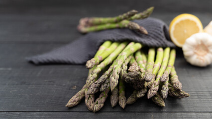 Close-up of green asparagus spears resting on dark wood with a deep blue kitchen towel, lemon and...