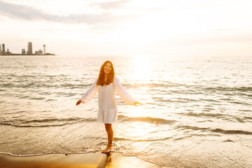 A joyful woman in a white dress has fun on the sunset beach. Young woman enjoying the sea and...