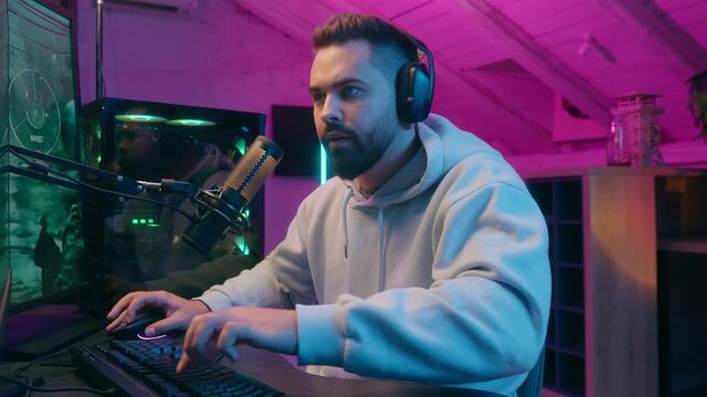 Addicted Caucasian guy male gamer streamer man play online battle enjoying smiling wearing headphones gaming at computer streaming in neon dark room at home match digital playing looking internet game