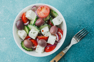 Greek salad with feta cheese in white bowl and fork on blue background