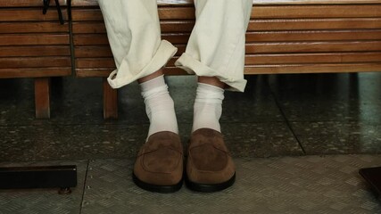 A person sits on a wooden bench at a busy transit station. They wear stylish loafers paired with white socks and loose cream-colored trousers, The girl takes off her shoes. - Powered by Adobe
