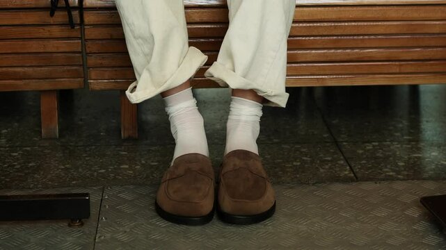 A person sits on a wooden bench at a busy transit station. They wear stylish loafers paired with white socks and loose cream-colored trousers, The girl takes off her shoes.